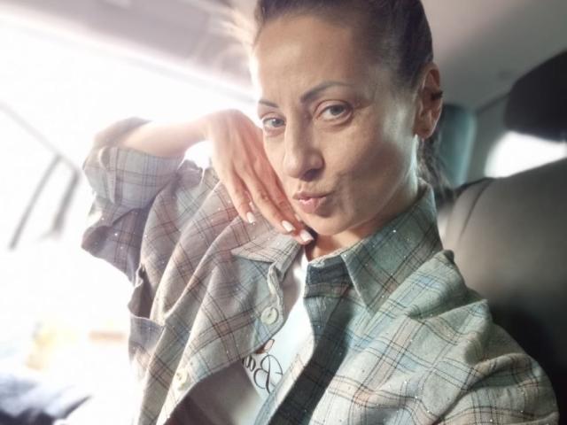 ViktoriiaLu with pensive look, hand on cheek, wearing plaid shirt, sitting in car, sunlight on face, subtle smile, dark hair in ponytail, white nails, relaxed vibe.