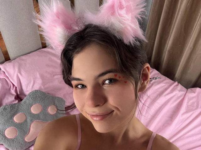 SwetHunny, con orejas de gato rosadas y peludas, sonriendo, tiene pequeñas marcas rojas en la mejilla, lleva una blusa rosa, está en una cama rosa con un cojín de gato gris..