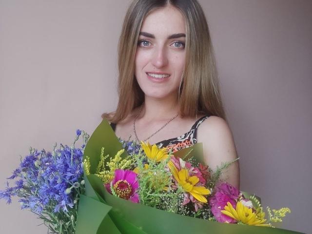 Feofaniia with long brown hair, blue eyes, and a smile, holding a colorful bouquet of flowers, including purple, yellow, and pink flowers, with green leaves.