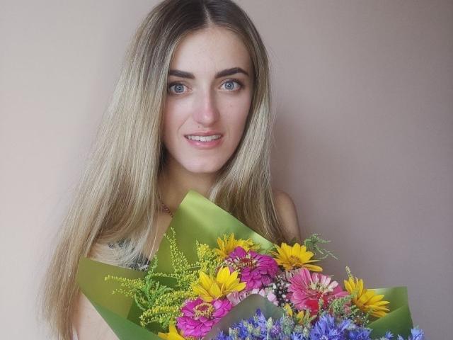 Feofaniia with long blonde hair, blue eyes, and a cute smile, holding a colorful bouquet of flowers. Her tits are partially visible, and she's wearing a green wrap.