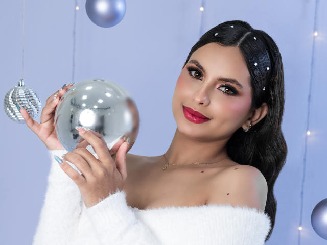 Dulce Coleman hält einen glänzenden silbernen Ball, trägt einen weißen, flauschigen Pullover mit einem Schulterteil, hat dunkles Haar, roten Lippenstift und ein süßes Lächeln und schaut in die Kamera..