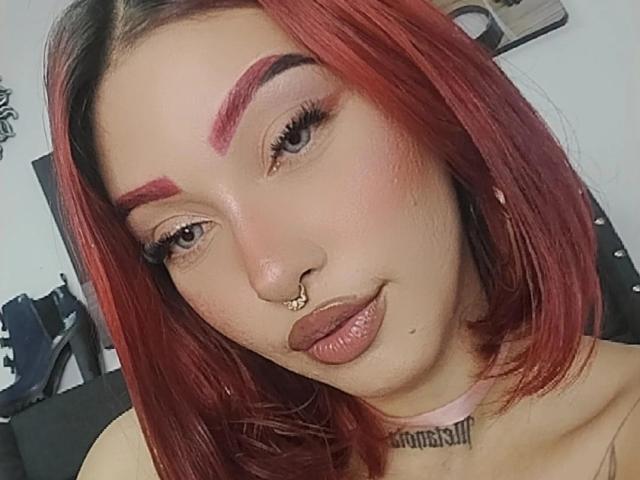 BriielaEva, con i capelli rossi, le sopracciglia rosa e un piercing al setto nasale, ha labbra marroni lucide e un collo tatuato, indossa una collana rosa e guarda la fotocamera..