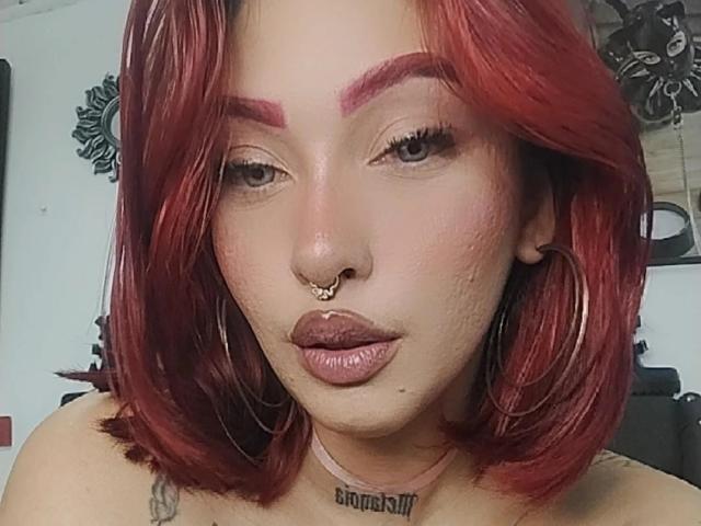 BriielaEva tem cabelo ruivo, sobrancelhas grossas e batom escuro. Ela está usando brincos grandes, um piercing no nariz e um piercing no septo, e tem tatuagens visíveis no peito..