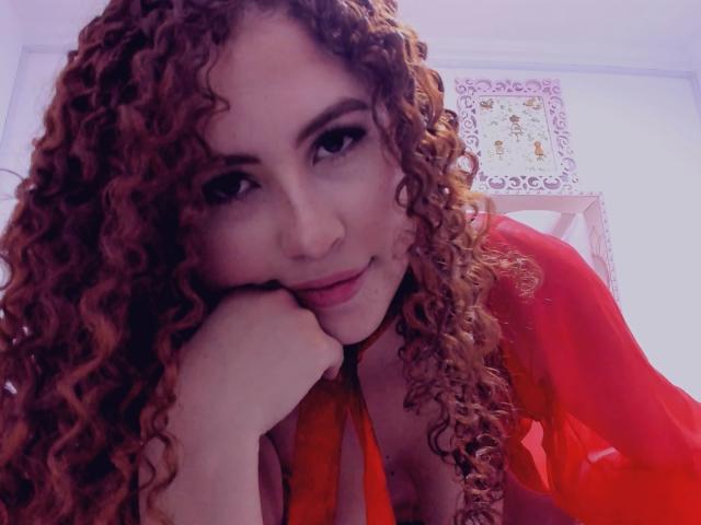 IrisDalton with curly red hair, red dress, and orange ribbon, resting her head on her hand, smiling slightly, showing her tits and red lips.