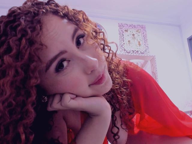 IrisDalton with curly red hair, head resting on hand, wearing a red top, looking at the camera with a slight smile, showing her tits, in a softly lit room.