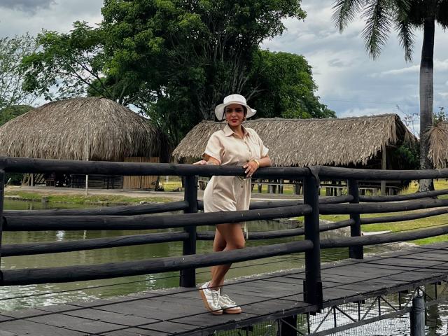 JessicaFiore, em pé em uma ponte de madeira, vestindo um vestido bege, um chapéu de sol branco e sandálias brancas, com o traseiro ligeiramente voltado para a câmera, apoiada em uma grade preta..