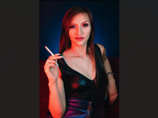 Aysha Smith mit langen, braunen Haaren, die ein schwarzes, glänzendes Kleid trägt, eine Zigarette in ihrer rechten Hand hält und mit pinkem Augen-Make-up in die Kamera schaut, bei roter und blauer Beleuchtung..