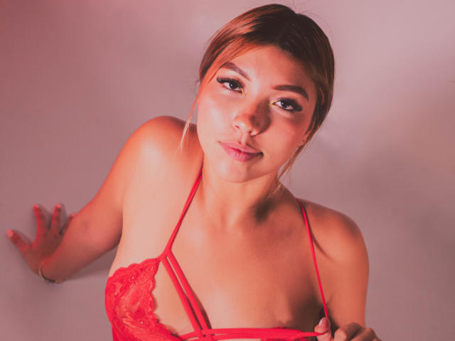 VaniaRizzo com um sutiã de renda vermelho, leve decote, e pele rosada, olhando para cima com um sorriso discreto, olhos castanhos, e cabelo castanho claro em um coque solto, segurando as alças do sutiã..