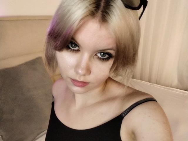 JaleesaLangworthy with blonde and purple-streaked hair, black tank top, blue eyes, and pink lipstick, looking up at the camera with a slight pout. Eyeglasses on her head.