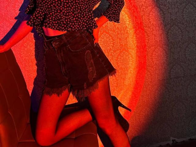 Seliina with a red sun-like glow on her, wearing a black floral top, frayed denim shorts, black high heels, and one knee up, standing against a textured wall.