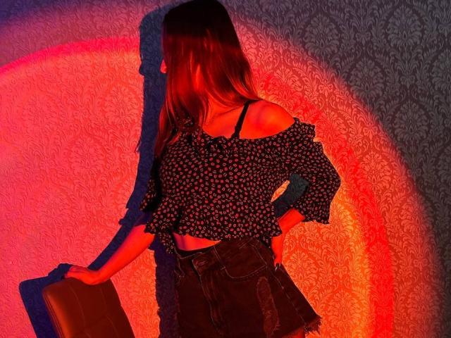Seliina with red and orange lighting, wearing a black off-shoulder floral top, exposing her tits, and dark denim shorts, holding a brown bag, shadow on the wall.