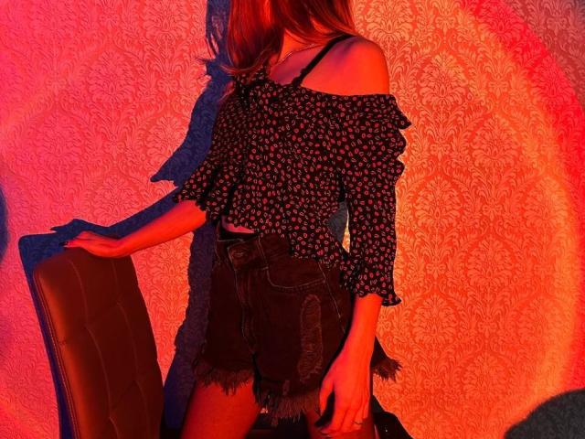 Seliina with long brown hair, off-shoulder black floral top, and frayed denim shorts, standing with arm on brown chair, illuminated by red and orange light.