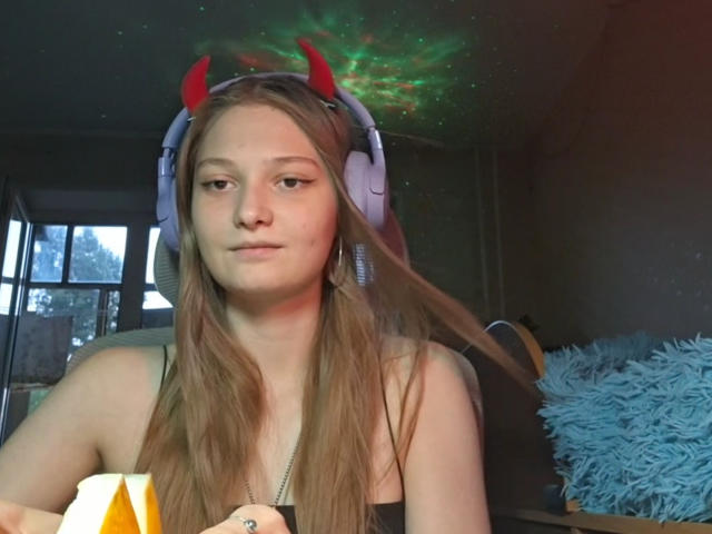 BrittanieManino with long brown hair, wearing red devil horns headphones, holding a donut, has a neutral expression, sitting in a dark room with a blue fluffy pillow.