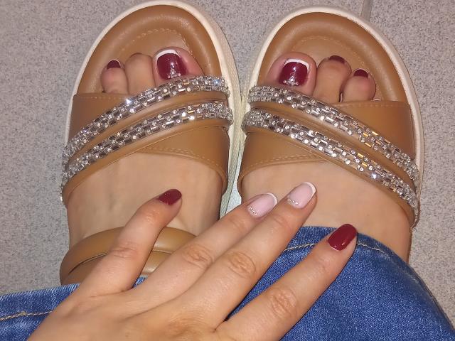 LindaMancini's feet with tan sandals, adorned with silver beads, showing red-painted toenails and pink nail with glitter on her index finger, resting on blue denim.