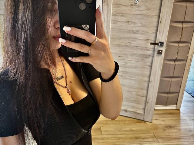 LilyMeeow with long brown hair, taking a mirror selfie. She's wearing a black zip-up top, showing her cleavage, with a necklace and ring. Her phone covers her face.