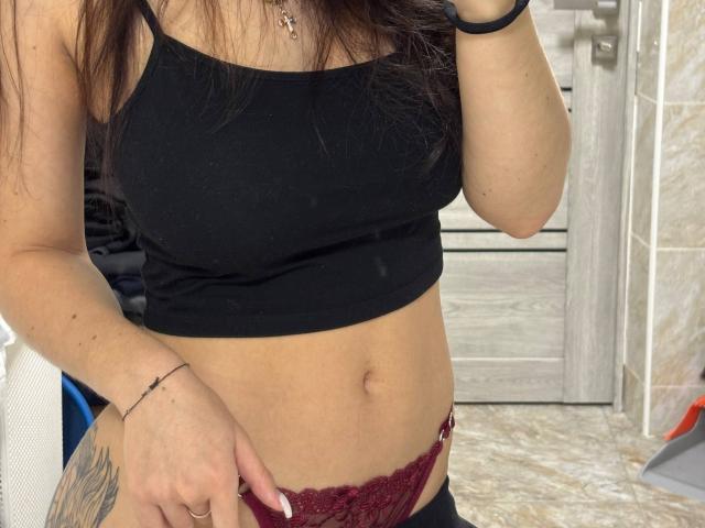 LilyMeeow's black tank top, showing her small tits, and red lace panties, with a tattoo on her right thigh and a bracelet on her left wrist, in a bathroom.