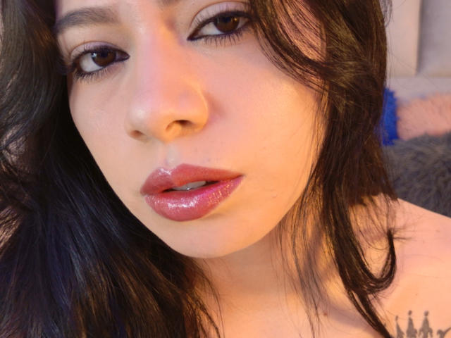 LindaMancini's face with shiny pink lips, dark wavy hair, and brown eyes. She has a slight tattoo on her right shoulder and a sexy, sultry expression.