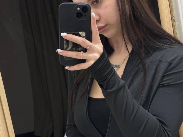LilyMeeow with long brown hair, black top, and jacket, holding a black phone with white nails, taking a mirror selfie, showing a tattoo on her chest.