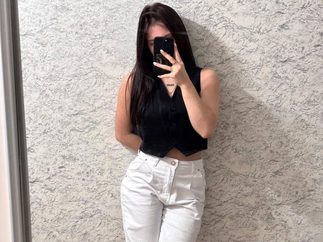 LilyMeeow with long dark hair, black sleeveless vest, and white high-waisted pants, holding a phone, covering her face, standing against a textured wall.