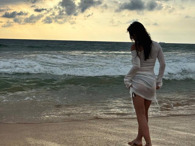 LilyMeeow with long dark hair, off-shoulder white dress, standing on beach, facing ocean, holding dress, showing bare ass, sunlight, and cloudy sky in the background.