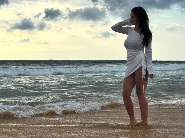 LilyMeeow with long dark hair, wearing a white sheer dress, stands on a beach, her hand on her head, her bare ass and thighs visible, waves in the background.