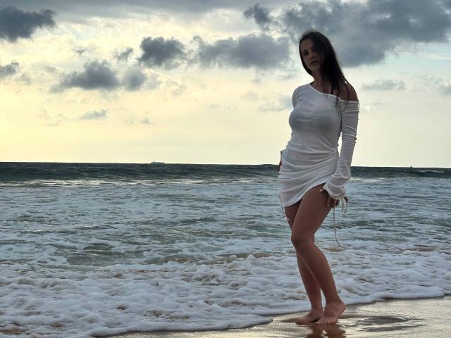 LilyMeeow stands on a beach, wearing a wet, white off-shoulder dress that clings to her tits and ass, with long dark hair, and a smile. Ocean waves behind.