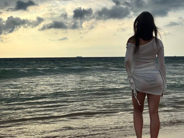 LilyMeeow stands on a beach, facing the ocean, in a sheer white dress, showing her ass, with dark wet hair and a cloudy sky above.