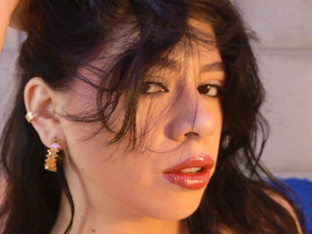 LindaMancini with messy black hair, glossy red lips, and gold earrings, looking to the side with a slightly open mouth. Her face is angled to the right.