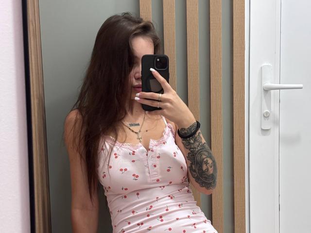 LilyMeeow with long brown hair, wearing a pink floral dress, holding a black phone, showing off her tits and tattooed arm, taking a mirror selfie.