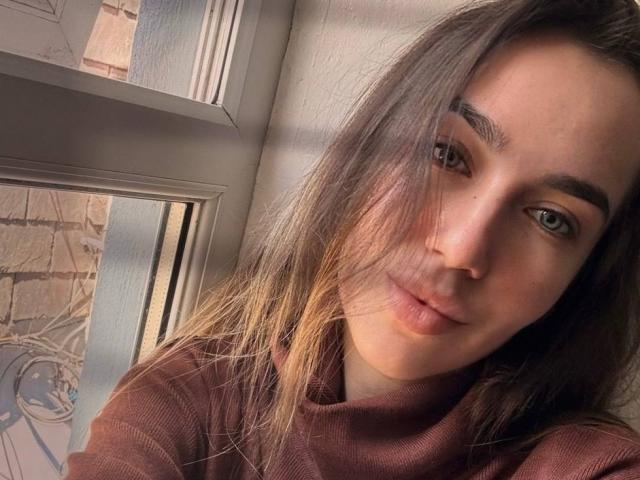 AraiaSheri with long brown hair, green eyes, and a subtle smile, wearing a brown turtleneck, sitting by a sunlit window. Her face is partially shadowed by her hair.