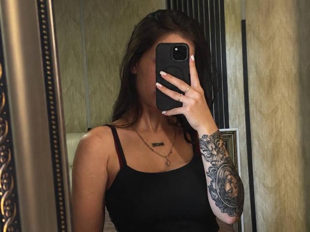 LilyMeeow with dark brown hair, black tank top, showing a tattooed arm, holding a phone covering her face, reflection in a mirror, gold necklace, red bra strap visible.
