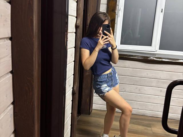 LilyMeeow with long brown hair, wearing a blue T-shirt and frayed denim shorts, taking a mirror selfie, showing her ass and small, round tits, in a wooden room.