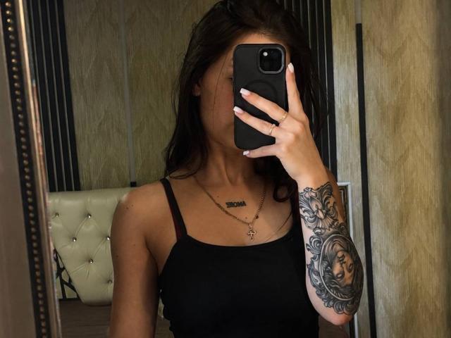 LilyMeeow taking a mirror selfie with a black phone, showing her black tank top, tattooed arm, and necklace. Her tits are visible, with her face mostly obscured.