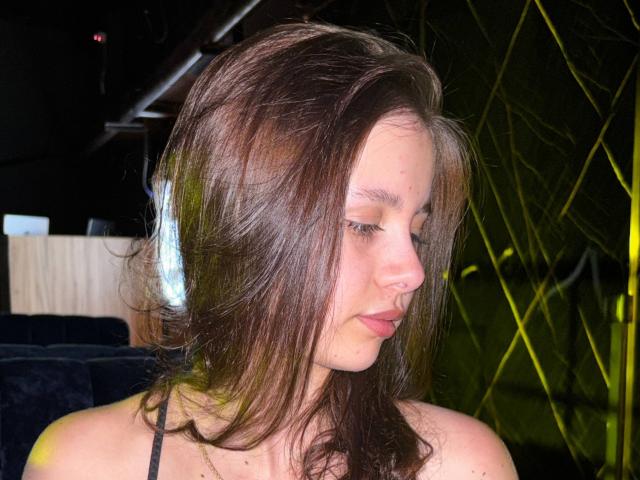 LilyMeeow has brown hair, a nose ring, and is wearing white headphones, looking down with a slight pout. She's in a dark, dimly lit room.