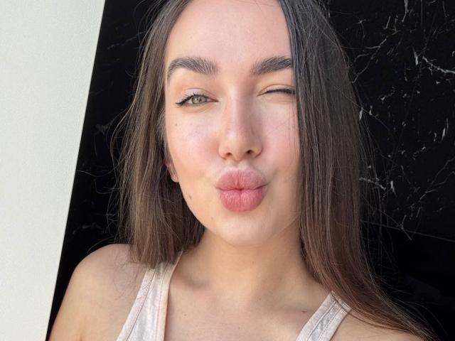 AraiaSheri has puffy pink lips, winks one green eye, and has long brown hair. She's wearing a white tank top, showing her small tits.