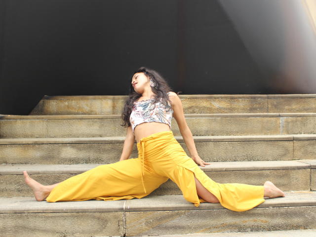 LucyySky with long black hair, tan skin, small tits, wearing a floral crop top and yellow pants, split-lying on stone steps, head tilted back, barefoot, looking relaxed.