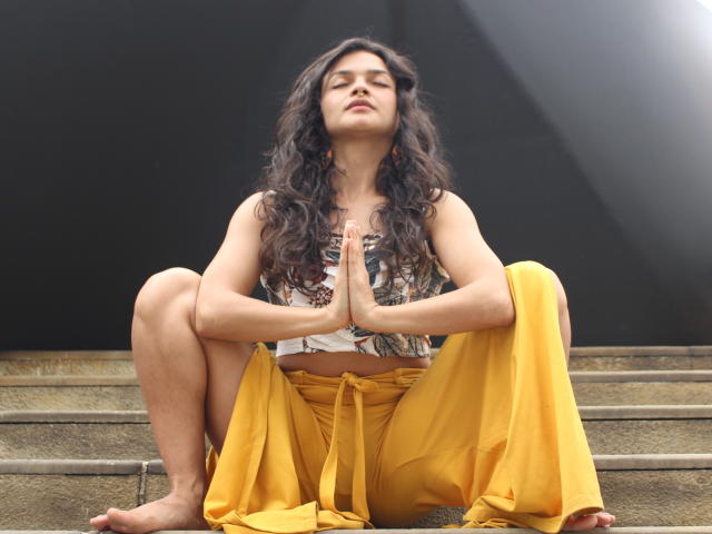 LucyySky with long curly dark hair, tan skin, and small tits, sits barefoot on concrete steps, legs spread, yellow pants, white patterned top, hands in prayer position, looking up.