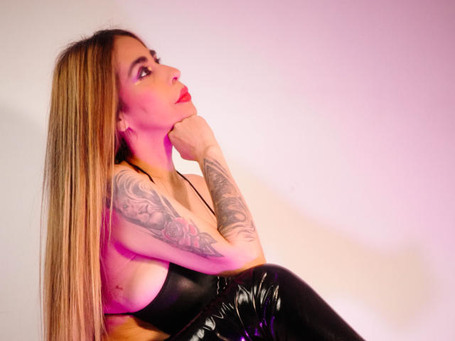 VictoriaJhonnes, con i suoi lunghi capelli biondi, il rossetto rosso, i pantaloni di pelle nera e i tatuaggi sul braccio, appoggia il mento sulla mano, guardando in alto con un'espressione seducente..