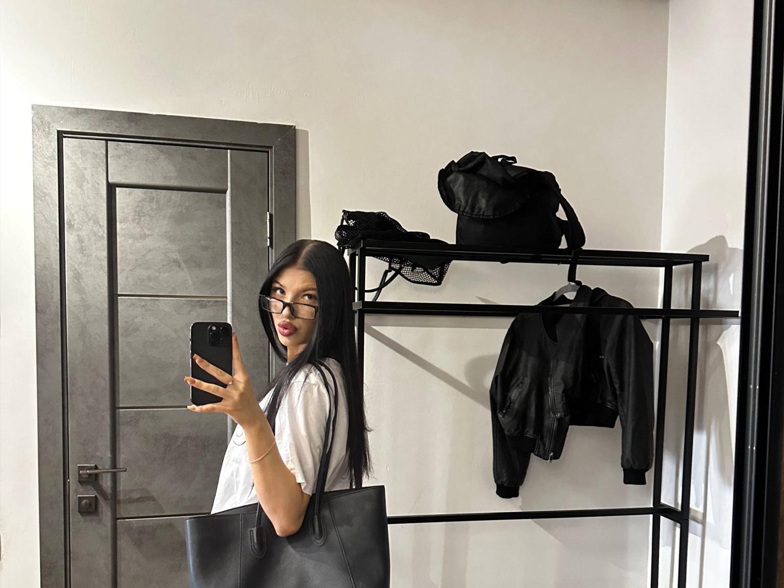 AngeliaHunger with long black hair, glasses, red lipstick, white shirt, black handbag, taking a mirror selfie, gray door, black shelf with bag and jacket, holding phone.