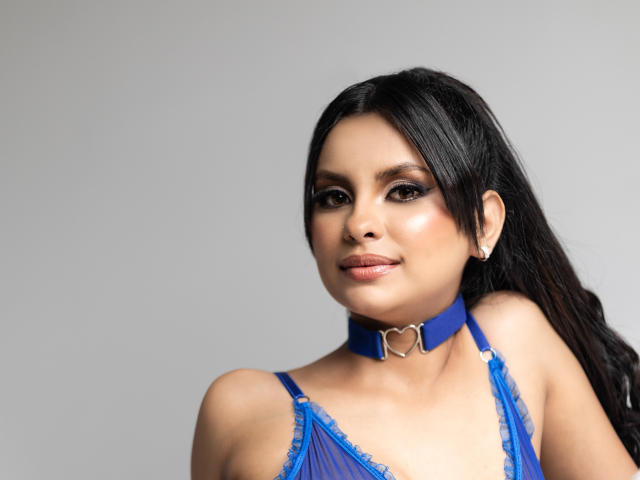 Dulce Coleman, con cabello oscuro, un sostén de encaje azul y un collar a juego con una hebilla en forma de corazón, sonriendo a la cámara. Sus pechos son pequeños y lleva un arete de plata..