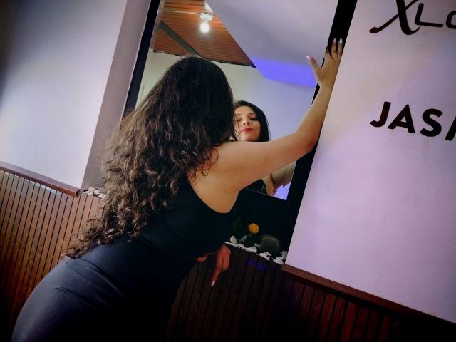 VelhMurphi with long curly brown hair, black backless dress, leaning against mirror, reflecting her face, showing off her ass, wooden wall paneling, blue ceiling light, looking at herself.
