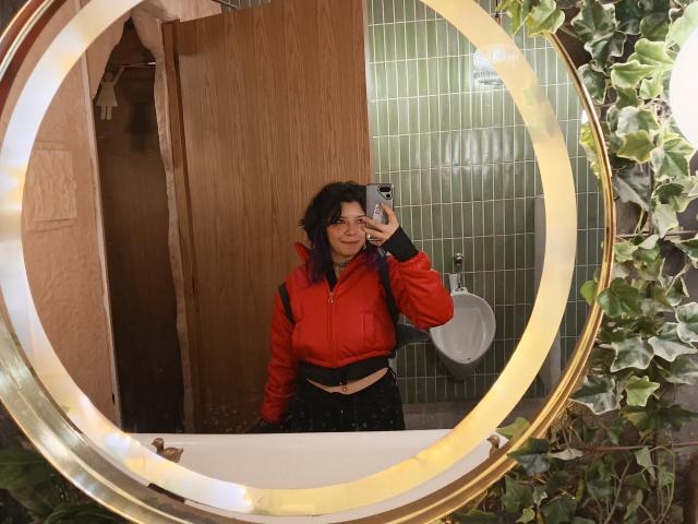 DarckVelvet with dark curly hair, red jacket, black skirt, holding phone in mirror, standing in front of urinal, surrounded by green tiles and vines, smiling.