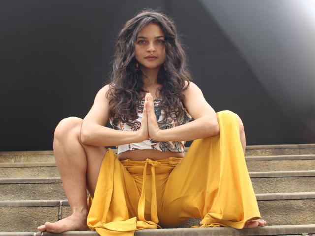 LucyySky with curly black hair, tan skin, and yellow pants, squatting with hands in prayer position, wearing a patterned top, barefoot, looking serious, on concrete steps.
