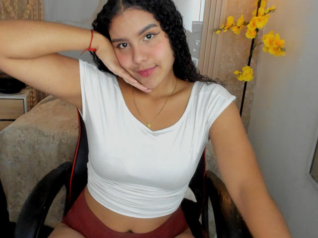 AriaVega, avec ses cheveux noirs bouclés, un crop top blanc moulant, un short rouge, assise sur une chaise, la main sur la joue, des fleurs jaunes en arrière-plan, regardant la caméra, avec un léger sourire..