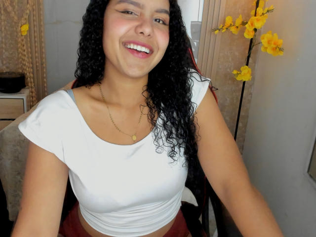 AriaVega with curly black hair, smiling, wearing a white crop top, showing her tits, and a gold necklace, seated, yellow flowers in the background.