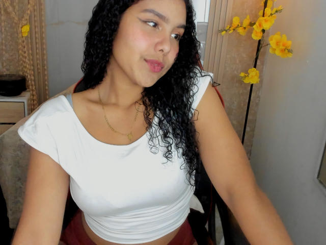 AriaVega with curly black hair, white crop top, and red skirt, wearing a gold necklace, sitting, pouting, yellow flowers in background. Her tits are small, she's cute.