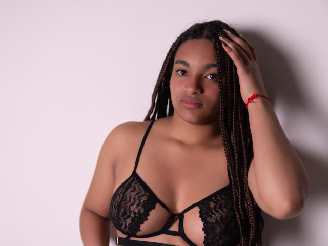 Kathe Palmer, con la pelle scura e i capelli intrecciati, indossa un reggiseno di pizzo nero, tocca la testa con un braccialetto rosso al polso, e guarda la fotocamera..