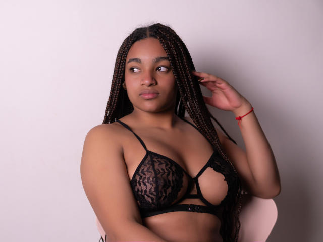 KathePalmer with long braided hair wearing a black lace bra with cutouts, showing her tits, has a red bracelet on her right wrist, looking to the side.