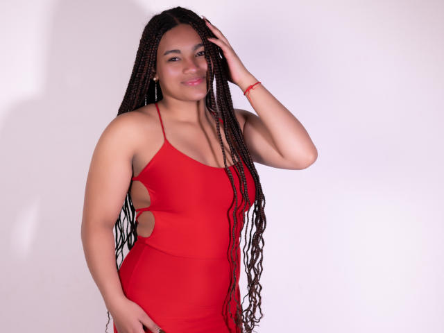 KathePalmer with long braided hair, wearing a red cutout dress, smiling, hand in hair, red bracelet, standing, white background, showing her tits and ass in the dress.