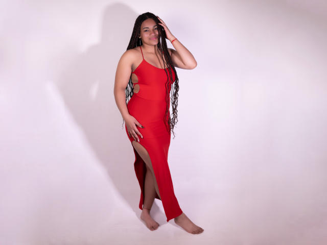 KathePalmer with long black braids, wearing a red dress with side slits, standing barefoot, hand in hair, red bracelet on wrist, smiling, full-figured, red dress covers tits.